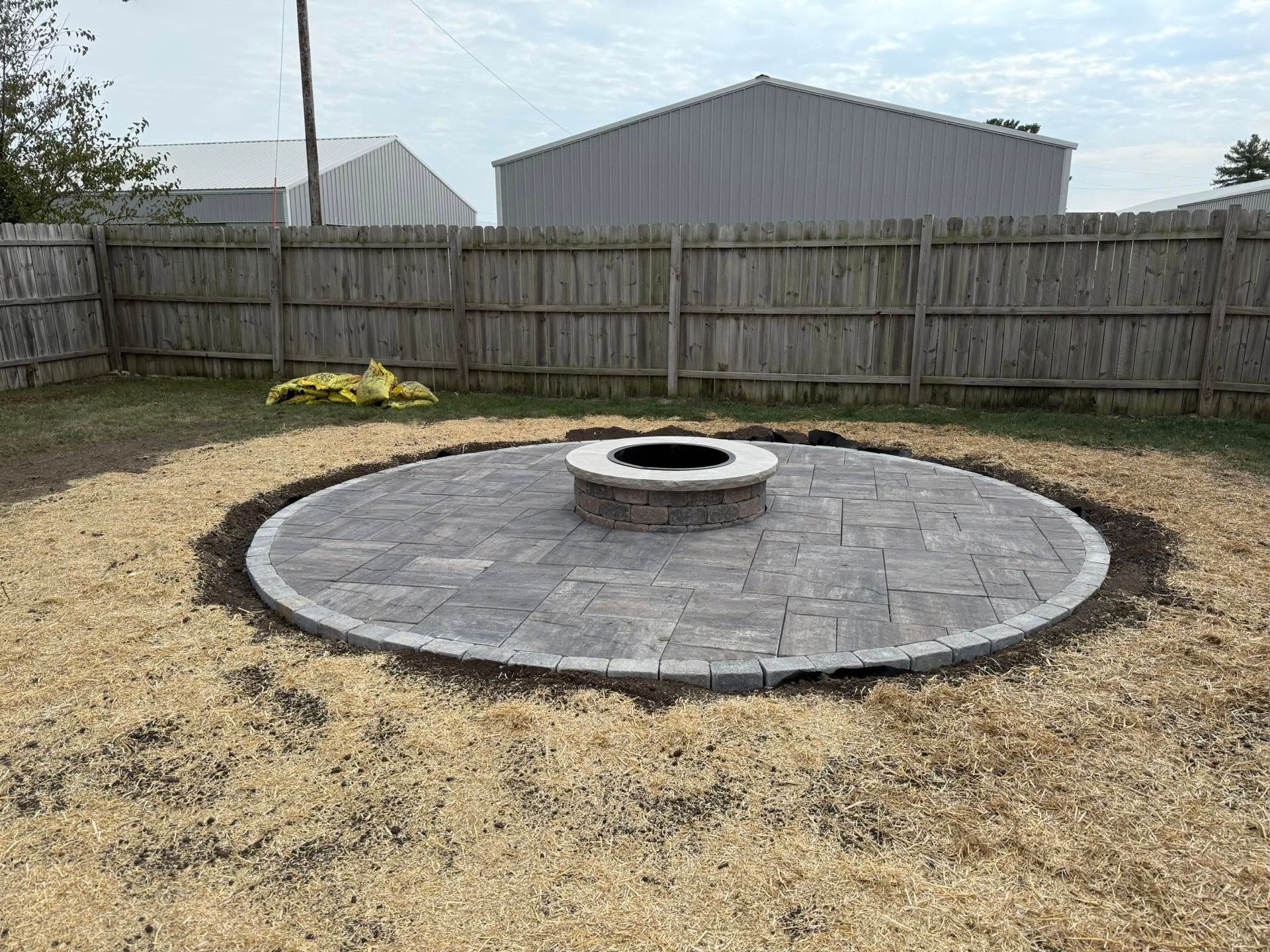 Backyard with a circular brick patio and central fire pit, surrounded by a wooden fence and dry grass.