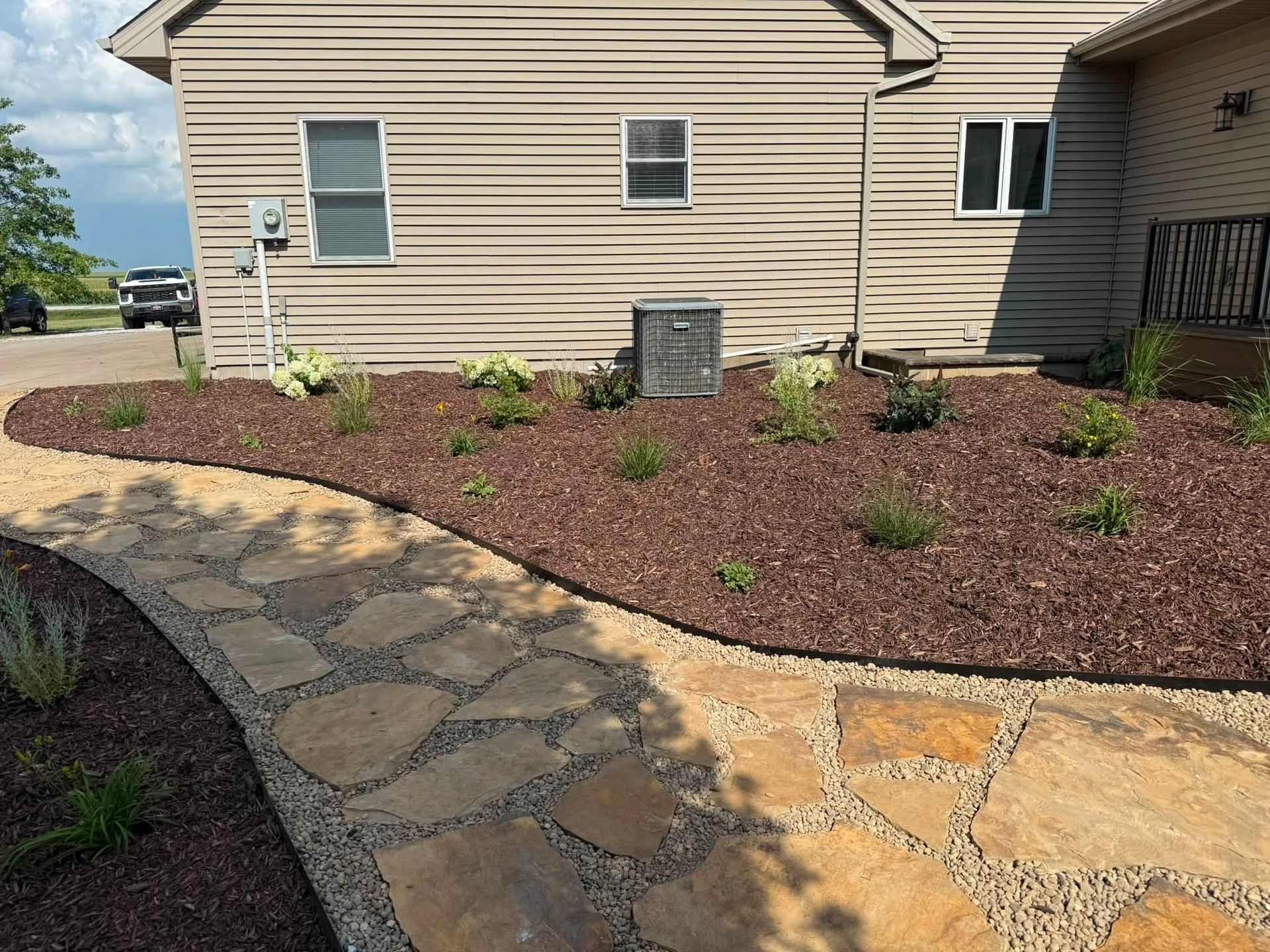 Stone walkway beside a beige house with a mulched garden bed and small shrubs