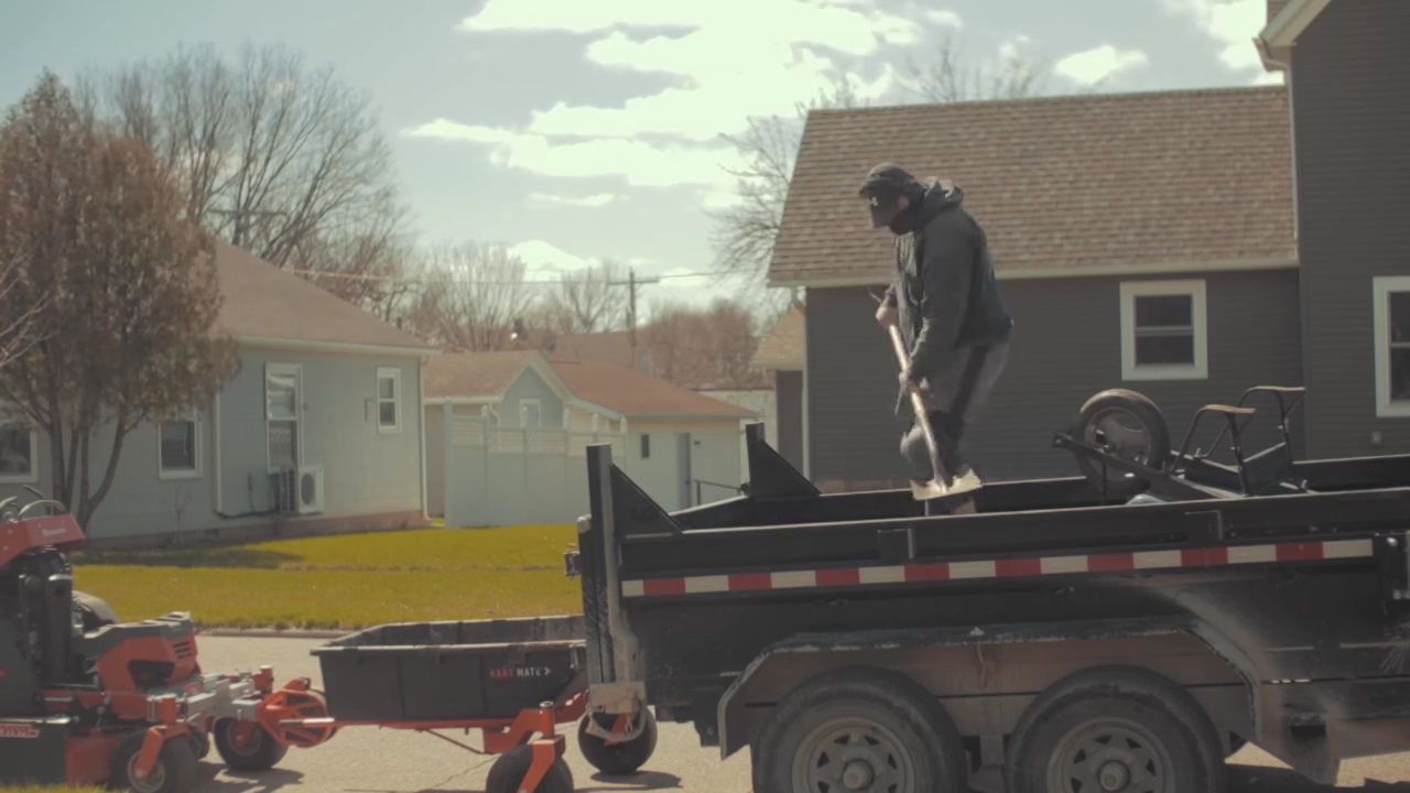 Person in dark clothing standing on a flatbed trailer in a suburban yard, with a tractor nearby.
