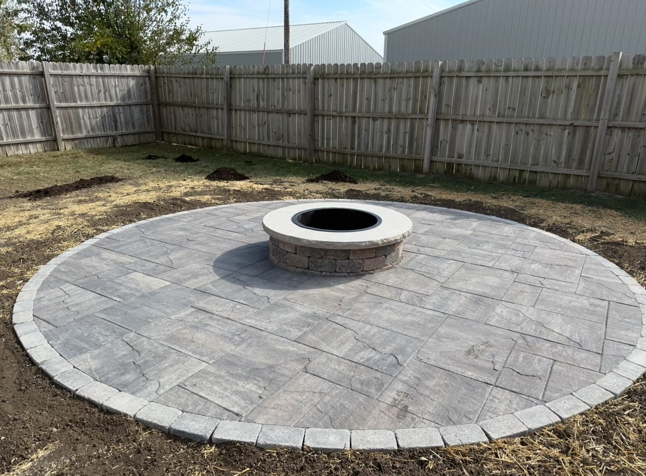 Circular stone fire pit patio in a fenced backyard with pavers and a grassy edge