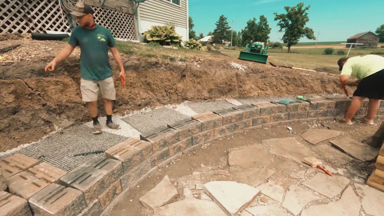 Workers installing brick pavers along a curved garden walkway beside a house.