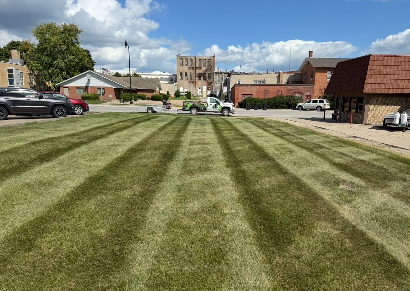 Striped lawn leading to a historic stone building and parked cars under a partly cloudy sky