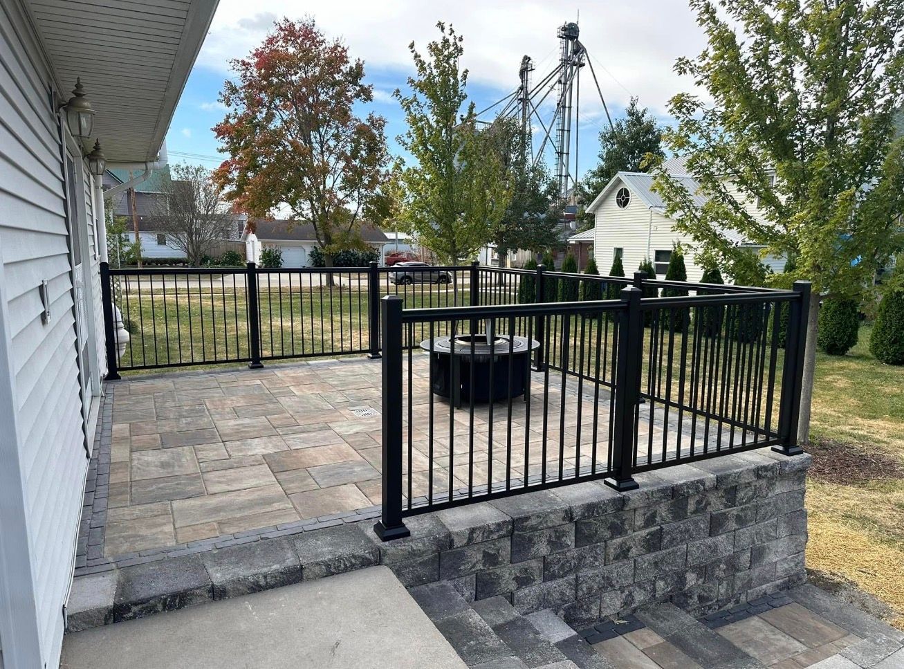 Black metal fenced patio with stone pavers, fire pit, and a view of trees and houses beyond