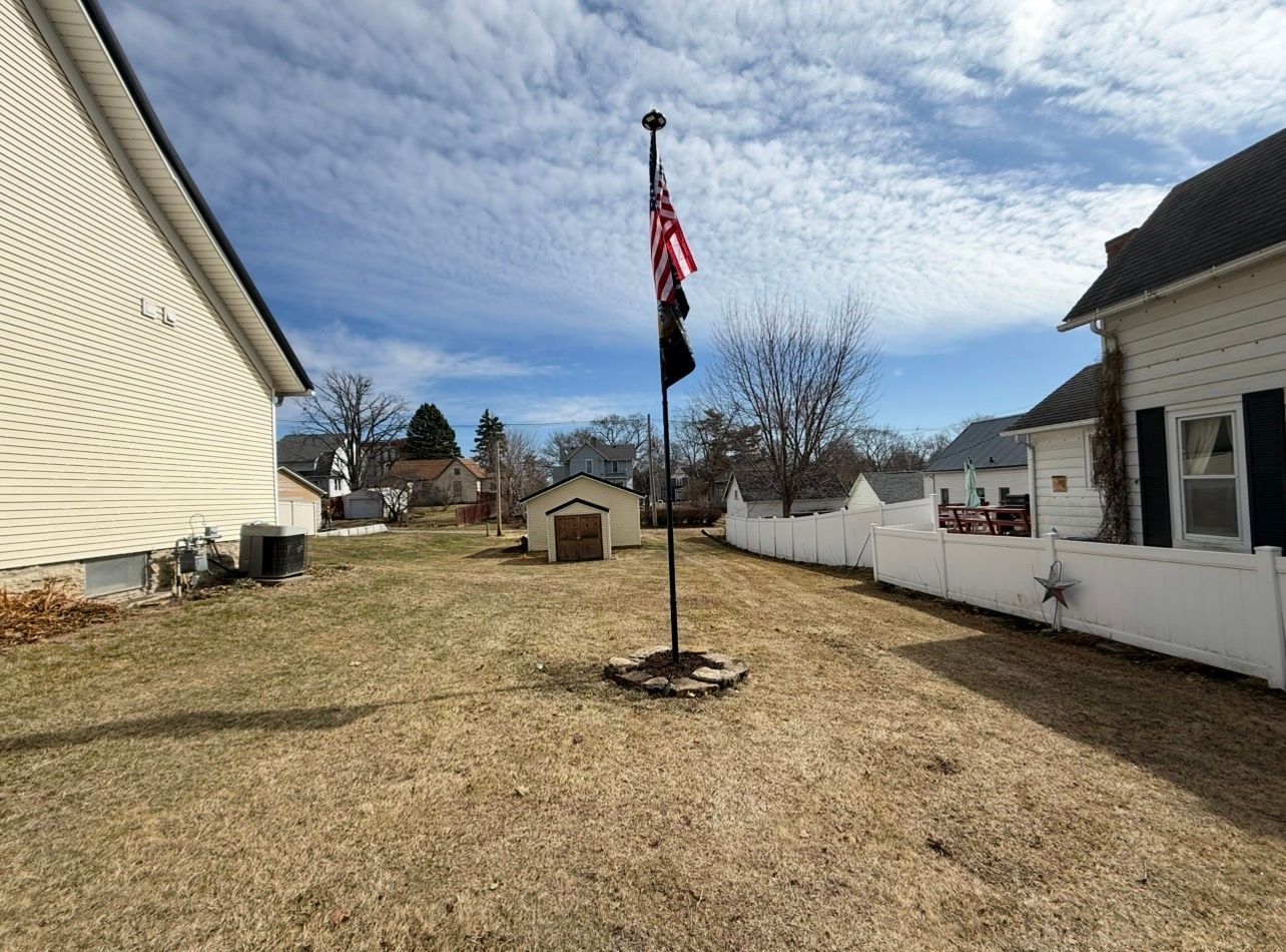 Backyard with a flagpole, winter-bare lawn, white fences, and houses under a partly cloudy sky