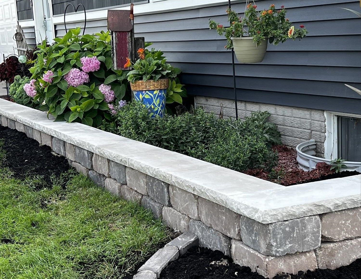 Stone retaining wall with blooming pink hydrangeas and potted plants beside a house exterior