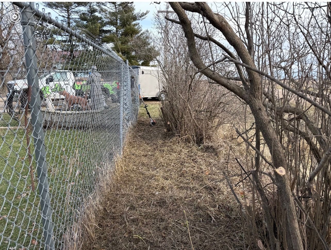 Leaf-covered narrow path beside a chain-link fence and bare trees, with a parked truck in the background