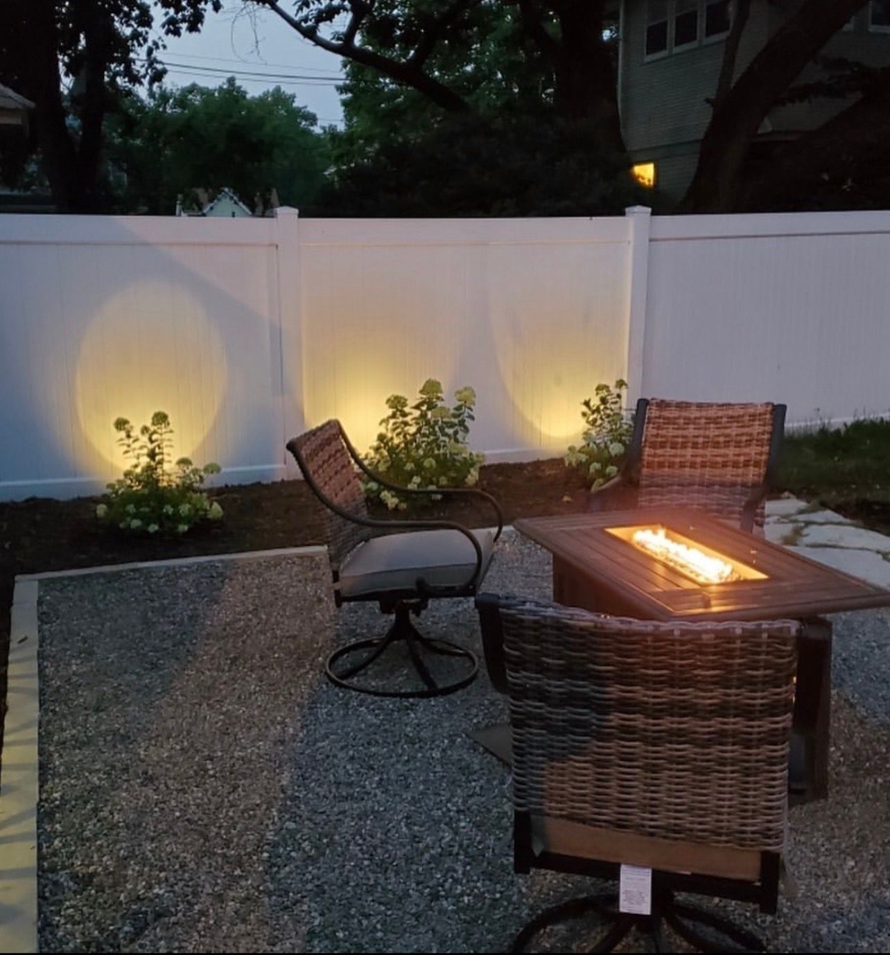 Backyard patio with wicker chairs around a rectangular fire pit at dusk, lit by warm landscape lights.