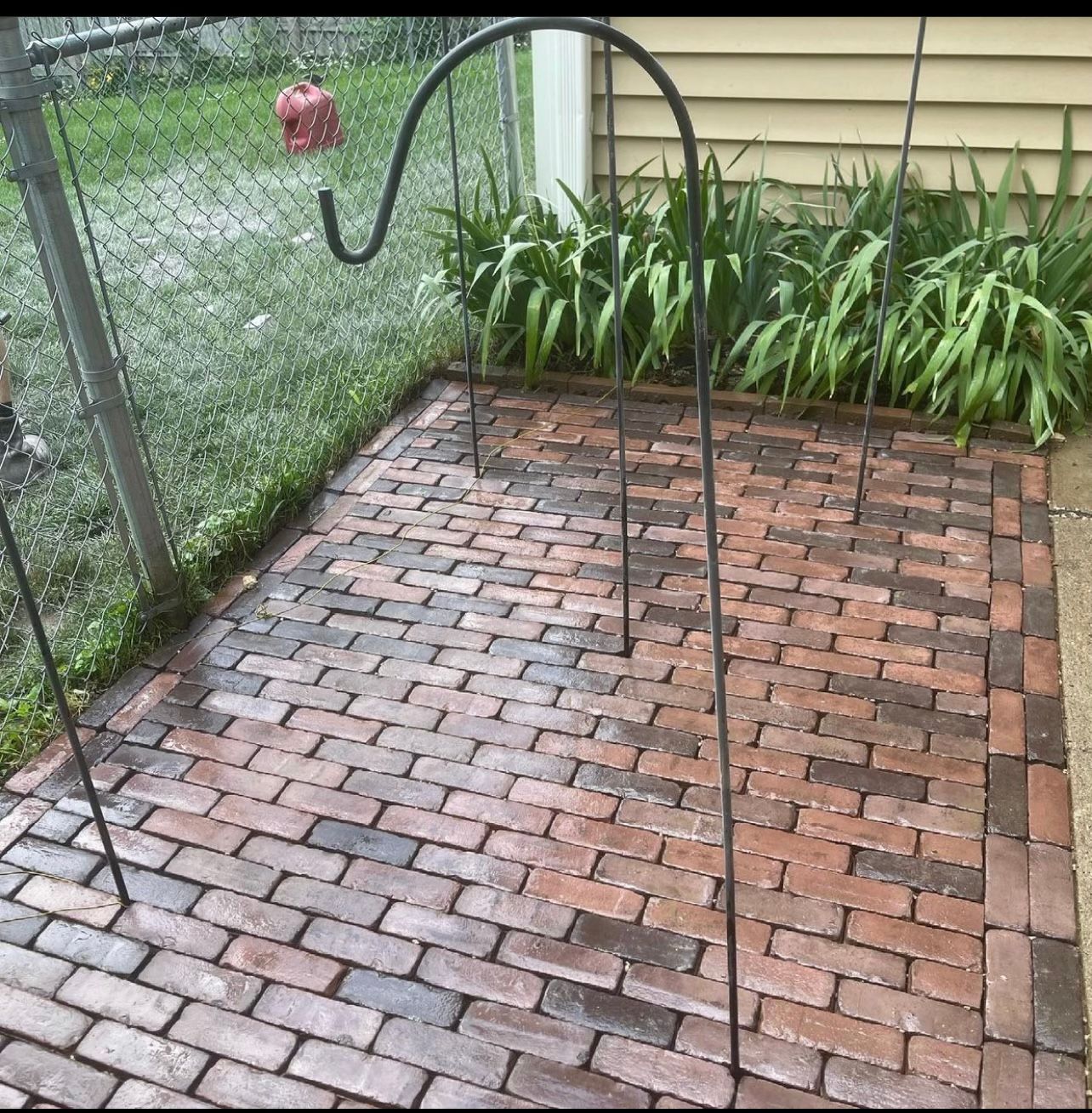 Brick patio with a hanging bird feeder beside a chain-link fence and garden bed.