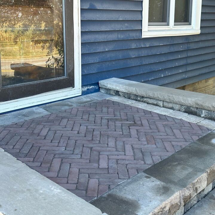 Brick paver patio beside a blue house with a window and concrete border