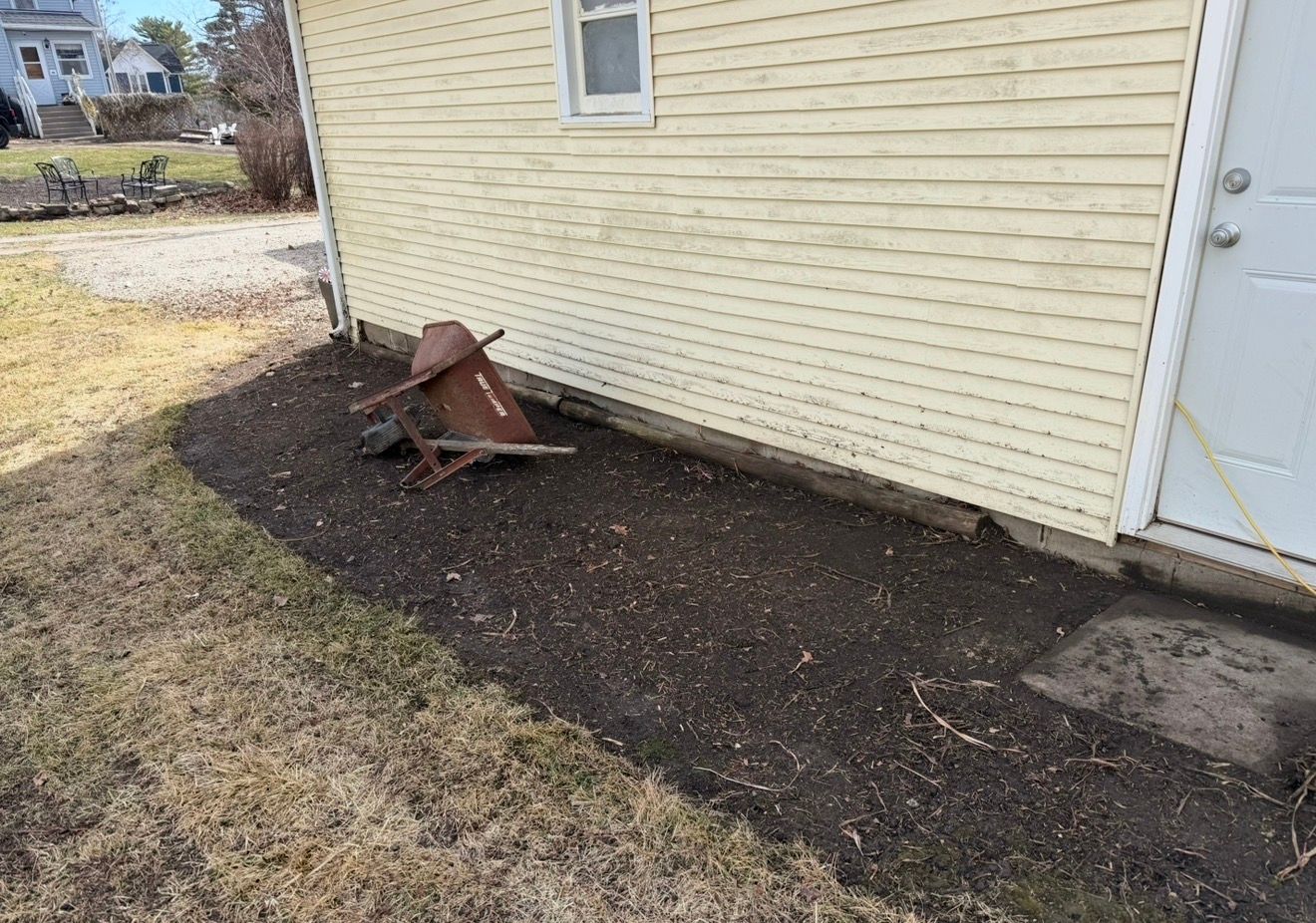 A rusted lawn mower lying on gravel beside a beige house wall and white door.