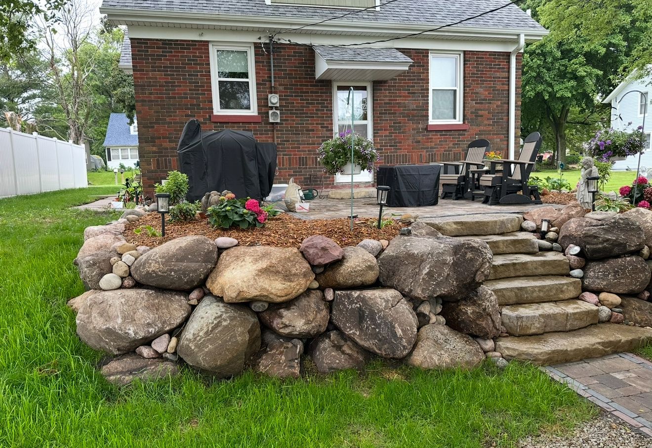 Brick house with a stone retaining wall, lawn, patio furniture, and outdoor grill in the backyard.