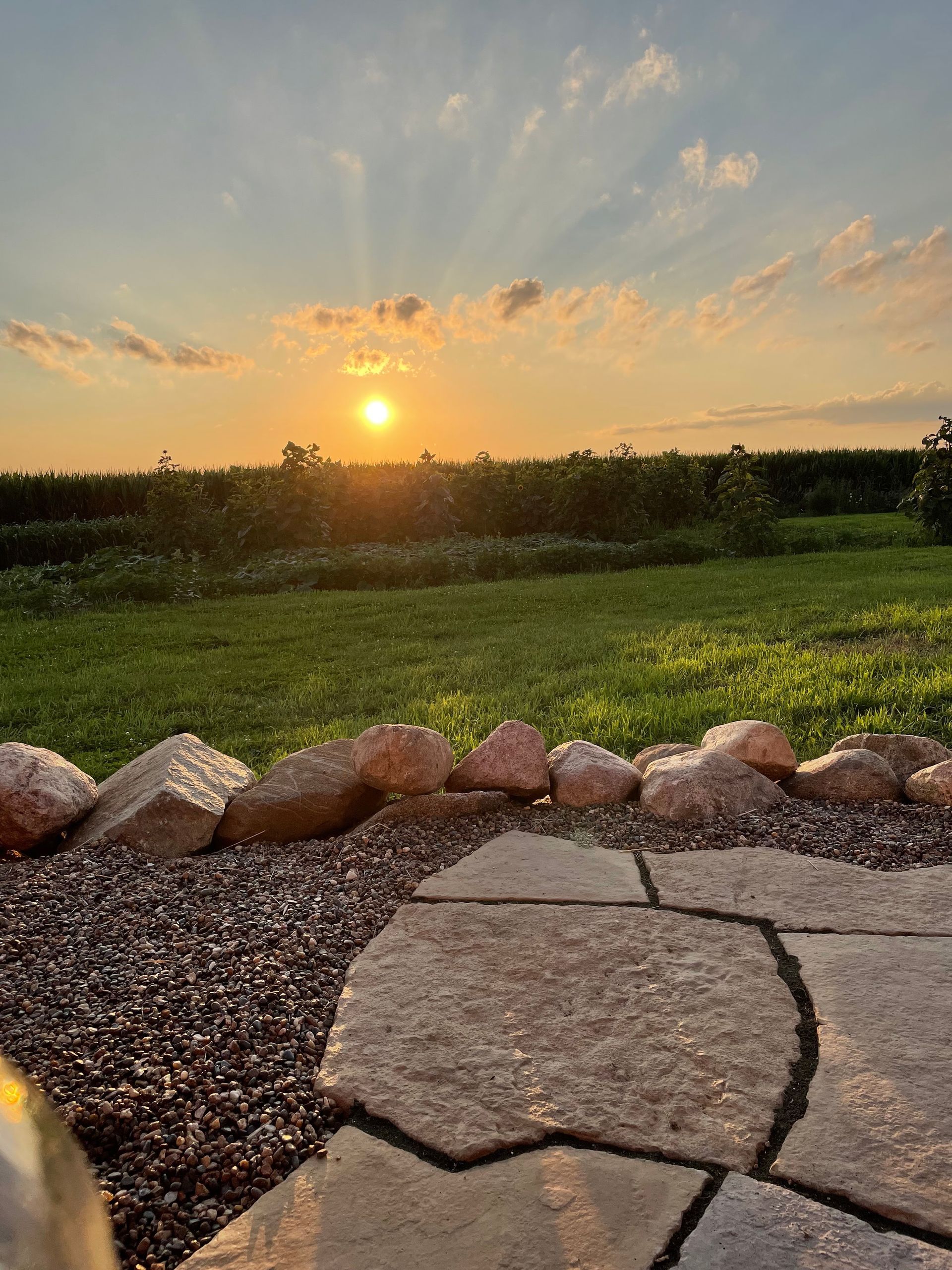 Sunset over a grassy field beyond a stone patio with rocks and scattered clouds