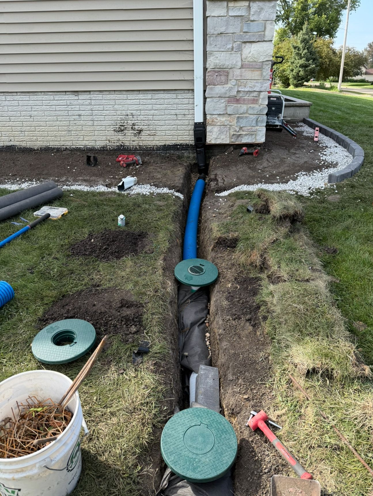 Digging trench beside house foundation with blue pipe and green utility covers in yard