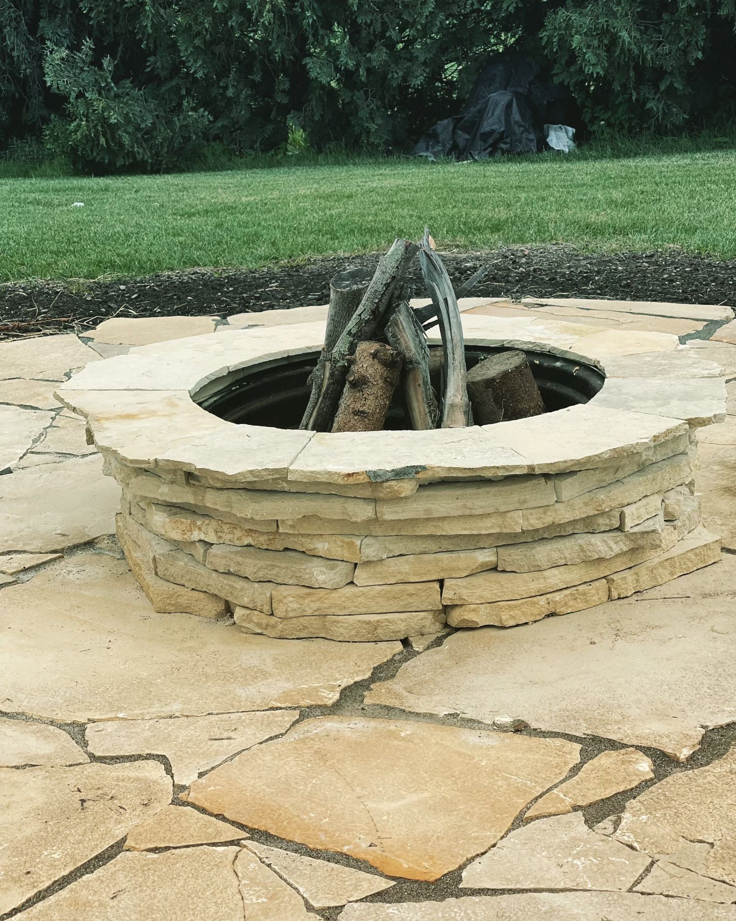 Stone fire pit with stacked rock rim on a paved patio, surrounded by grass and trees