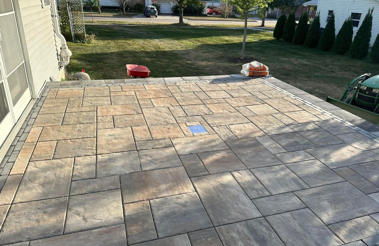 Stone patio under construction with a blue tape square, two tool bags, and a yard in the background
