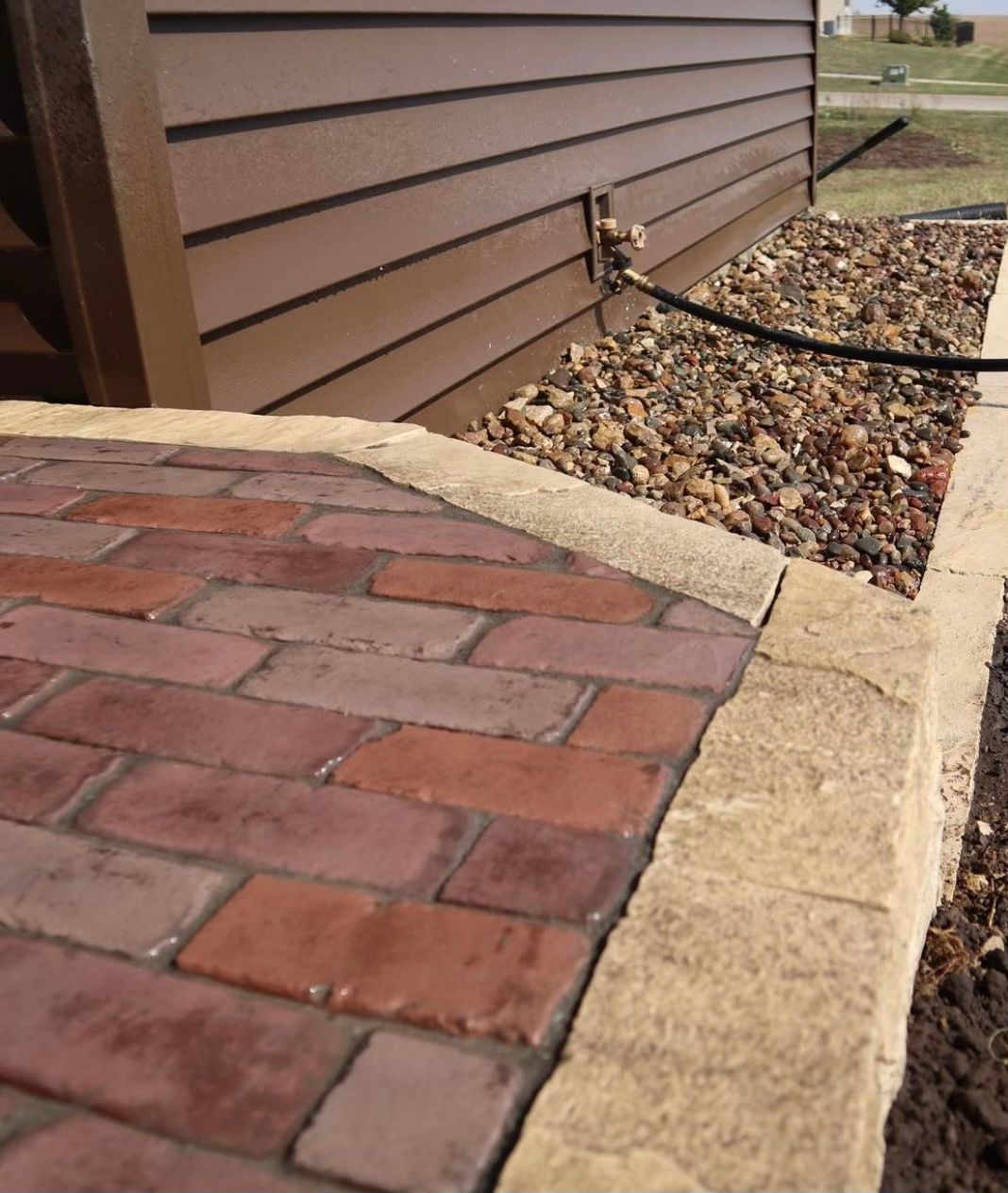 Brick and stone walkway beside a brown house with a gravel border and hose spigot