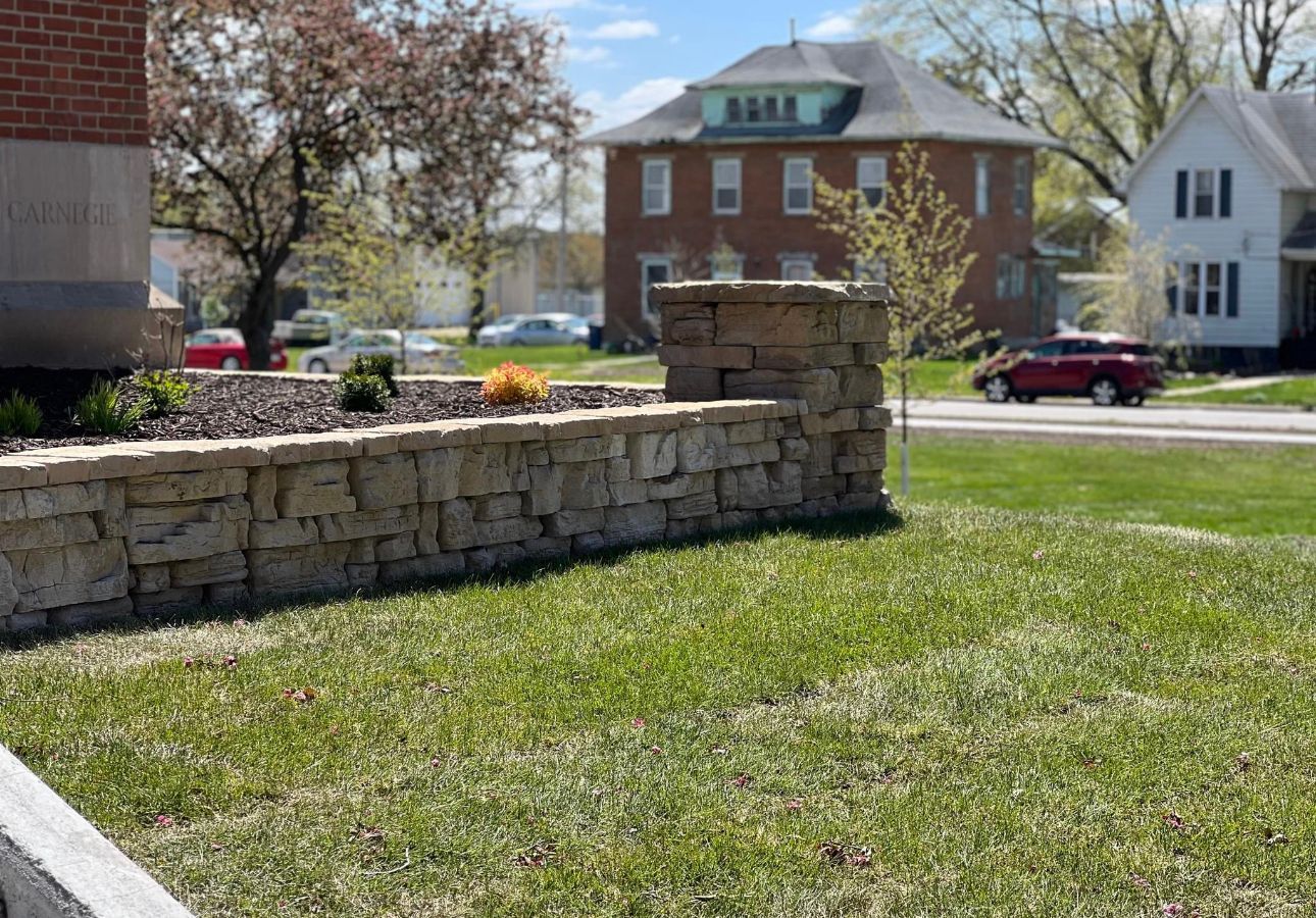 Stone retaining wall with flowers in front of a brick house on a sunny suburban street