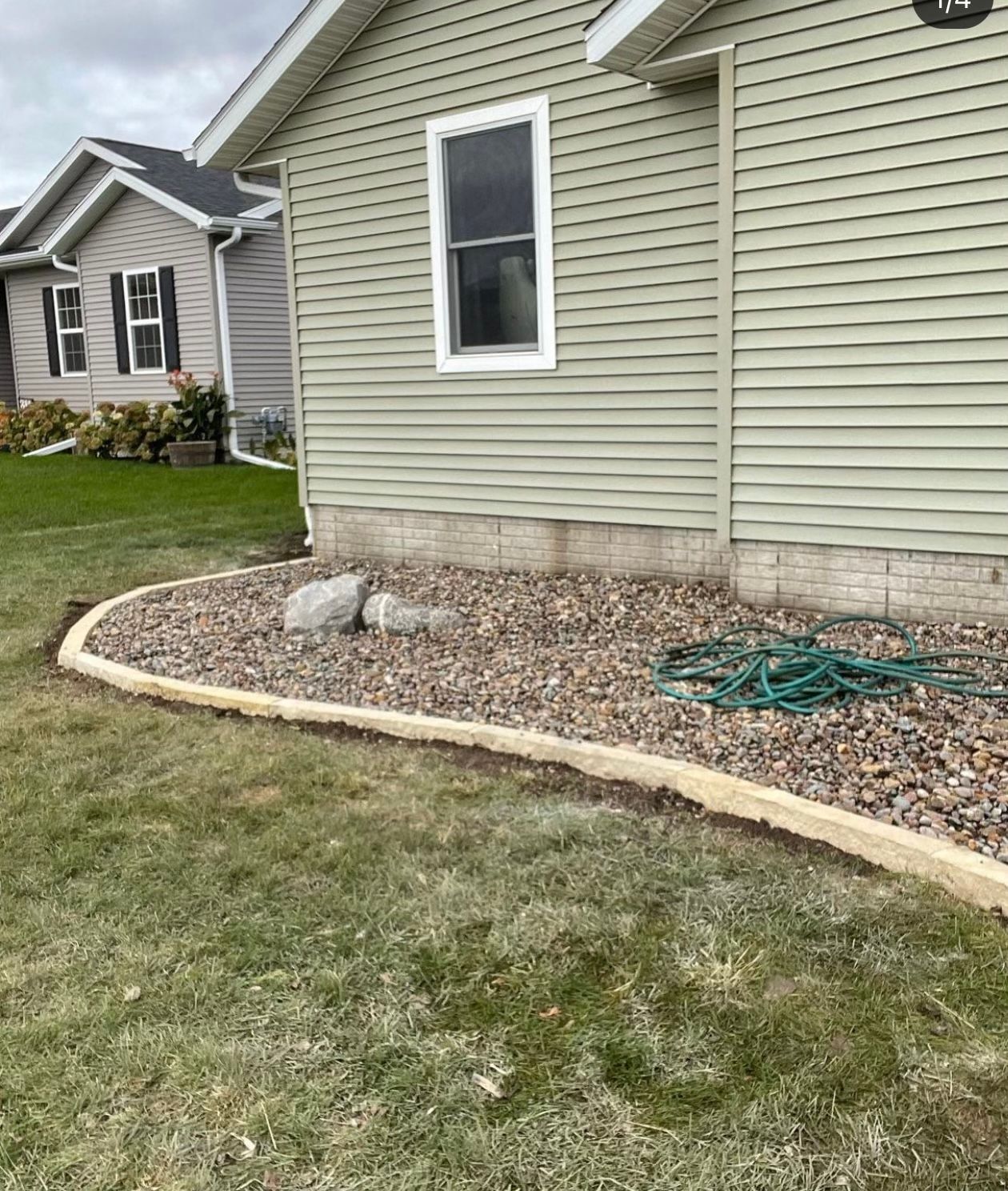 Side of a house with beige siding, small window, rock bed, curved edging, and a green hose on the ground.