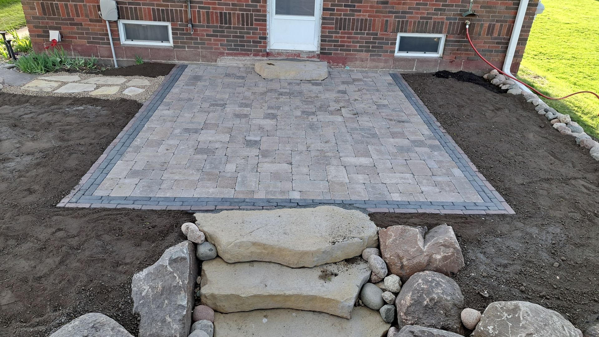 Backyard patio pavers bordered by soil, with stacked stones in front of a brick house.