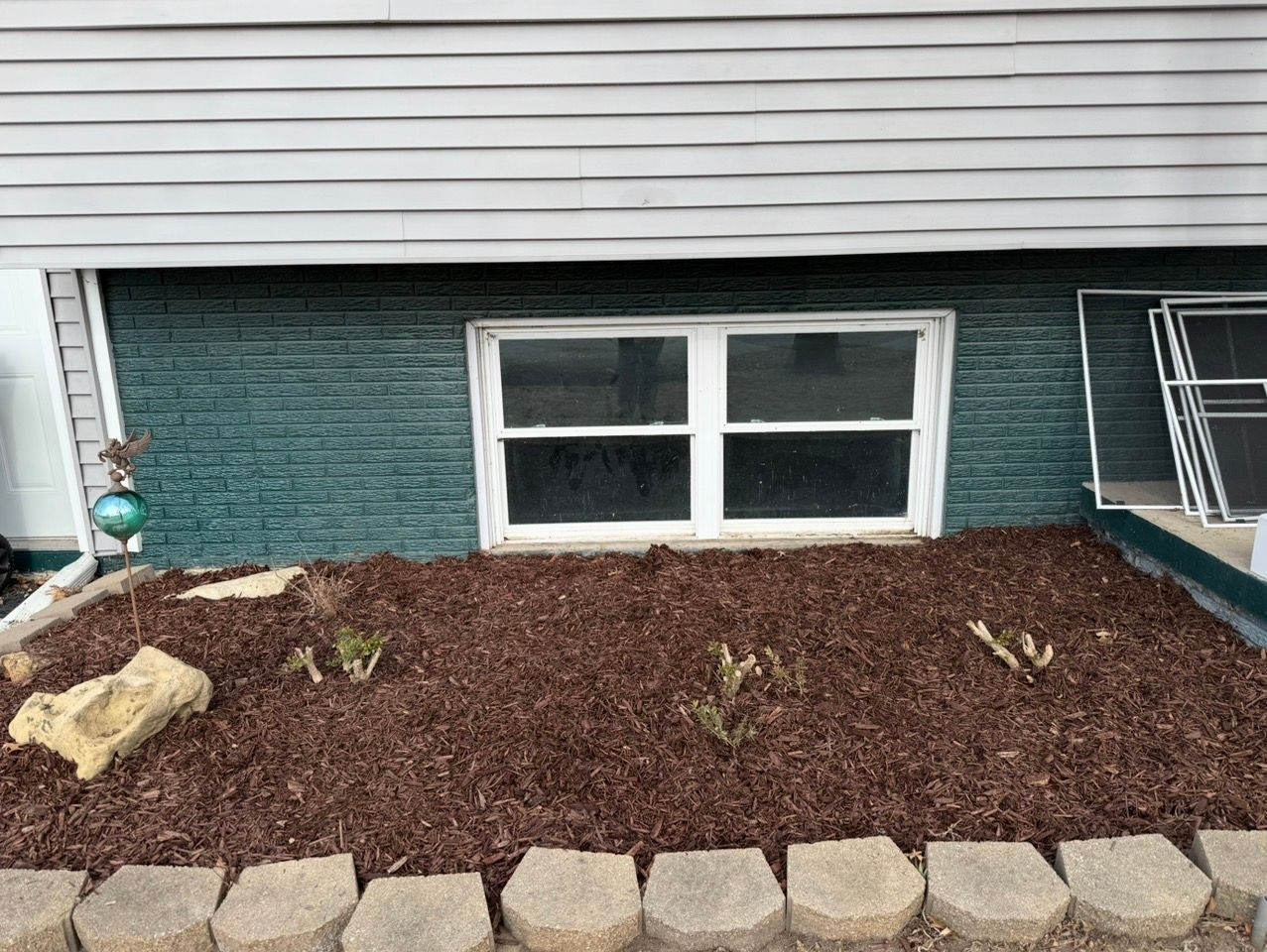 Front house window with brown mulch bed, stone border, and small plants under white siding