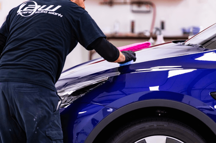 Man polishing a blue car in a garage, using a sponge with pink detailing tools.