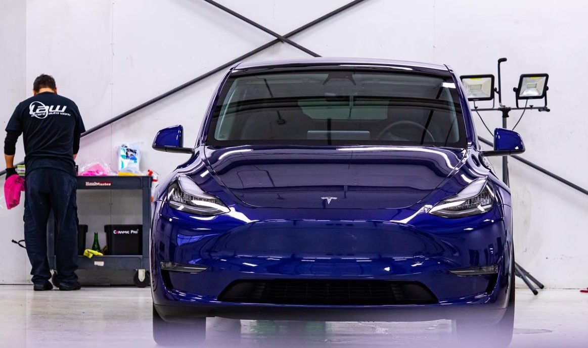 Person cleaning a blue Tesla Model Y in a garage, with detail lights and supplies nearby.