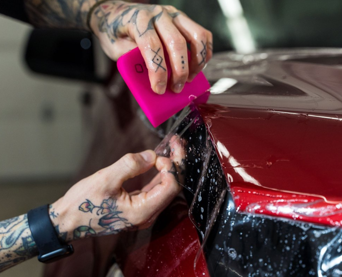 Person with tattooed arms applying protective film to a red car using a squeegee in a garage.