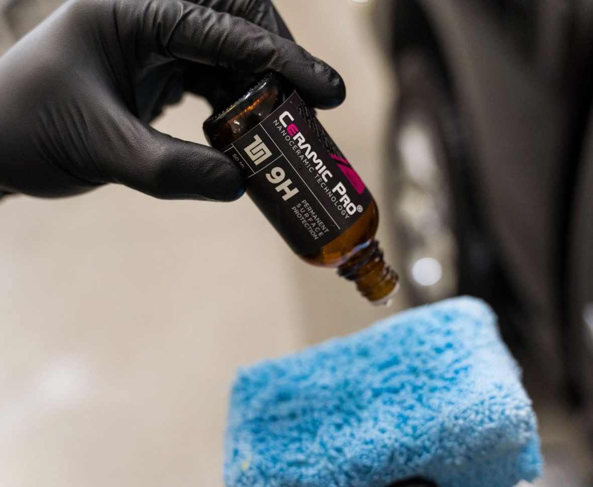 Black-gloved hand dispensing Ceramic Pro 9H onto a blue microfiber applicator. Automotive detailing.