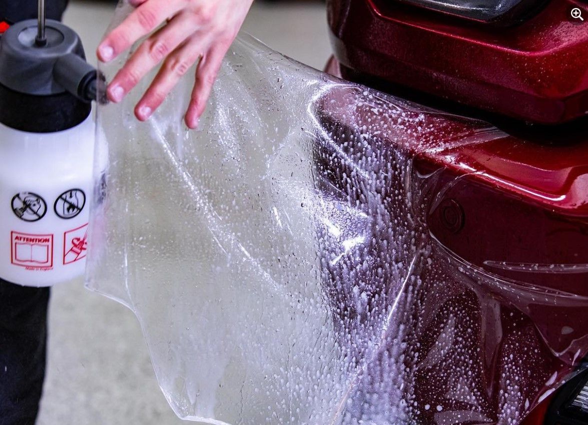 Person spraying soapy water onto a clear protective film on a red car.