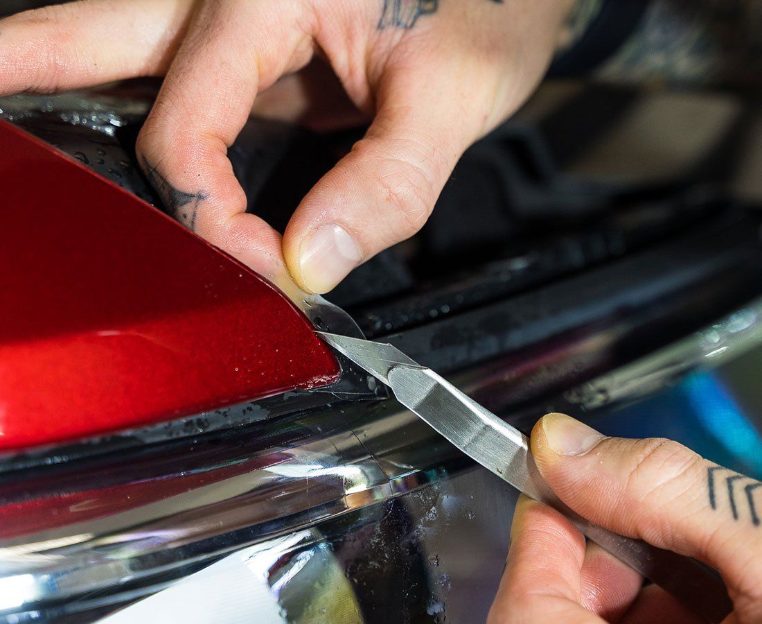 Hands using a blade to trim film on a red car part, next to chrome trim.