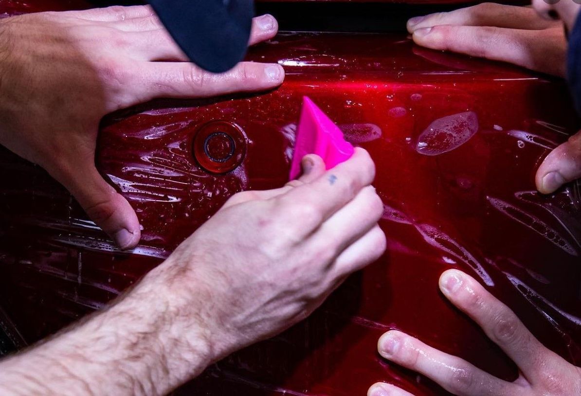 Hands applying protective film to a red car bumper, using a pink squeegee.