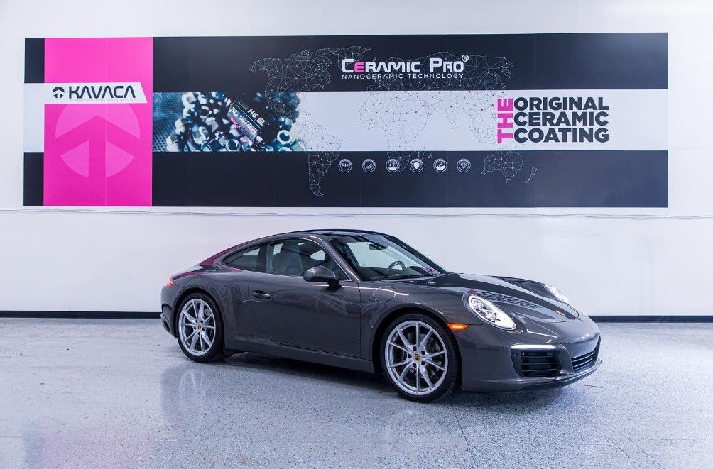 A gray porsche 911 carrera is parked in front of a ceramic coating sign.