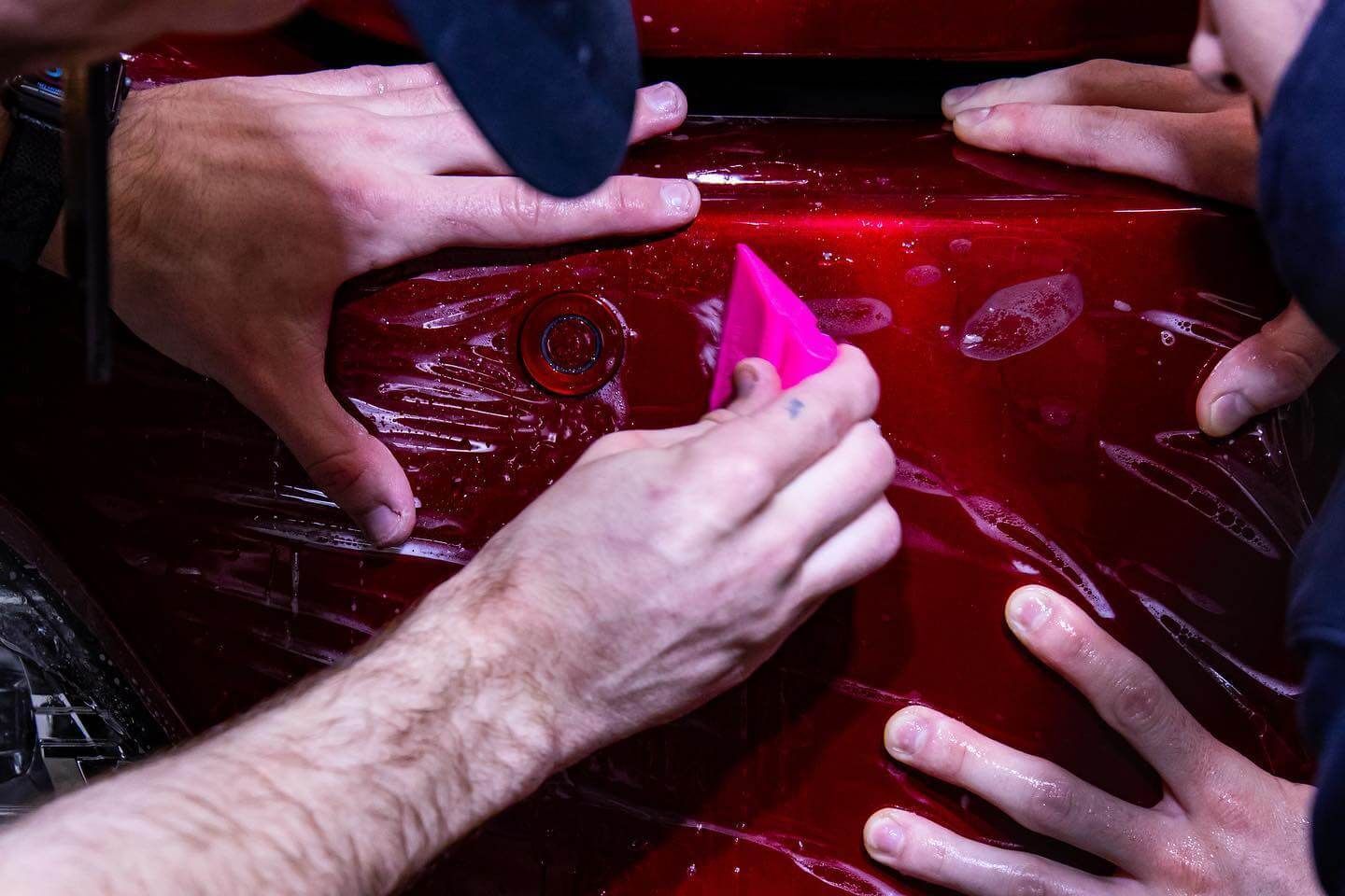 Hands applying a protective film to a red car bumper, using a pink squeegee to remove bubbles.