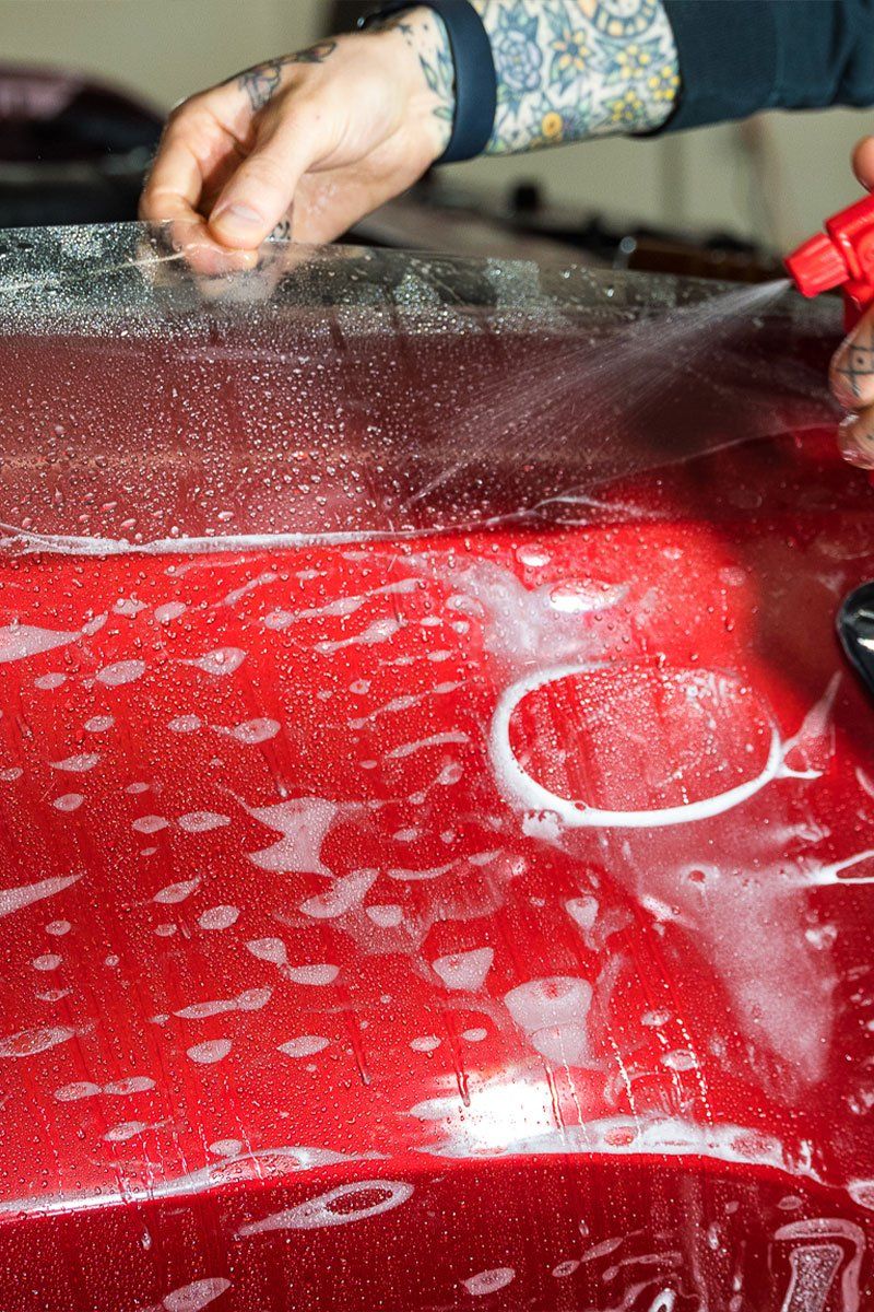 Person spraying soapy water on a red car surface before applying a clear protective film.