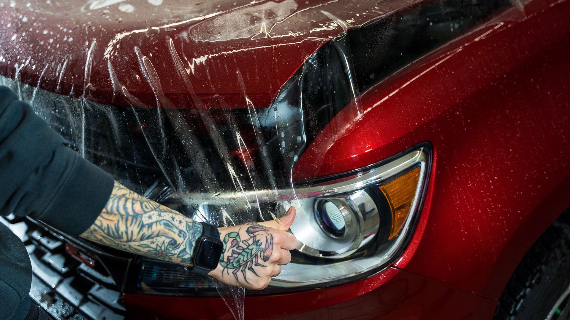 Person applying protective film to a red car headlight with tattoos visible on their arm.