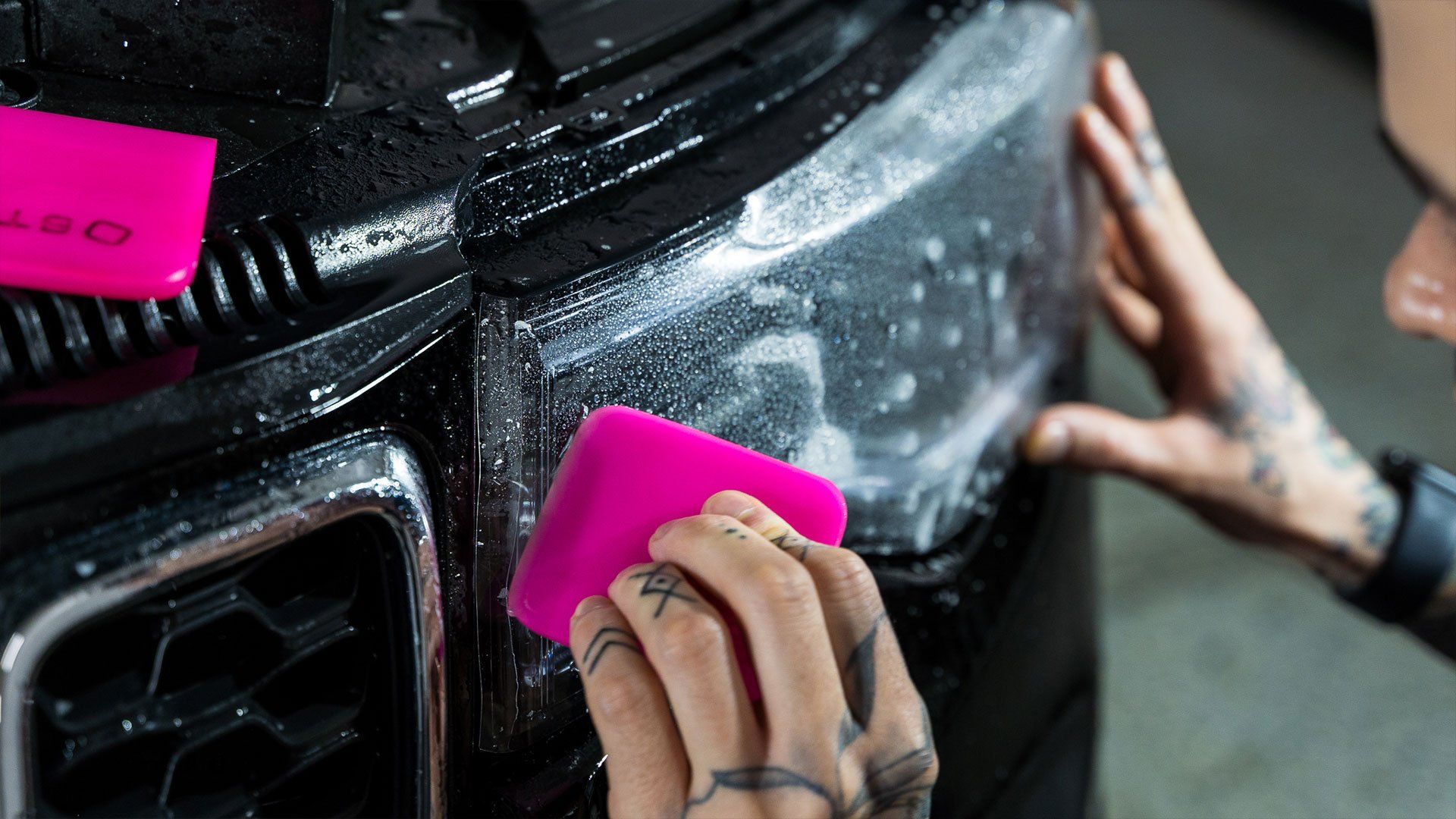 Person applying film to a car headlight using pink squeegees, detail shot of hands and car.