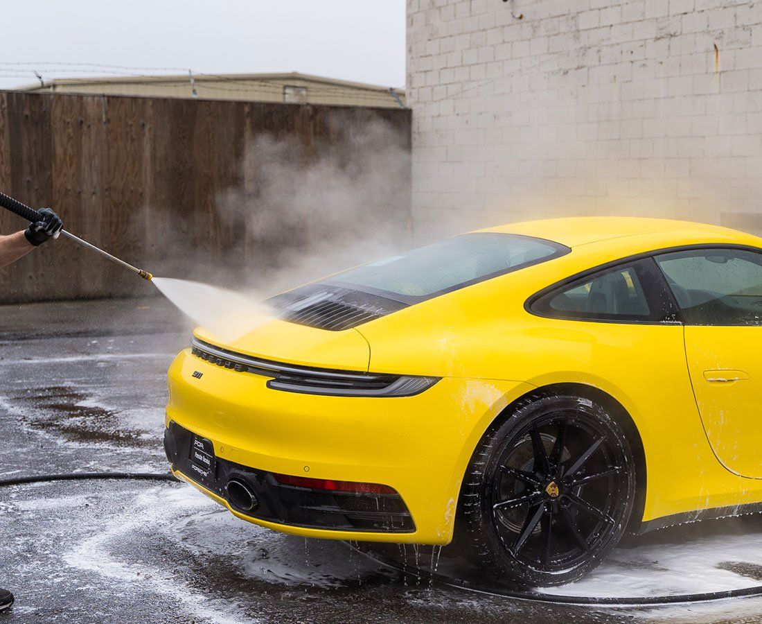 Yellow Porsche being washed with a pressure washer; foam and water are visible.