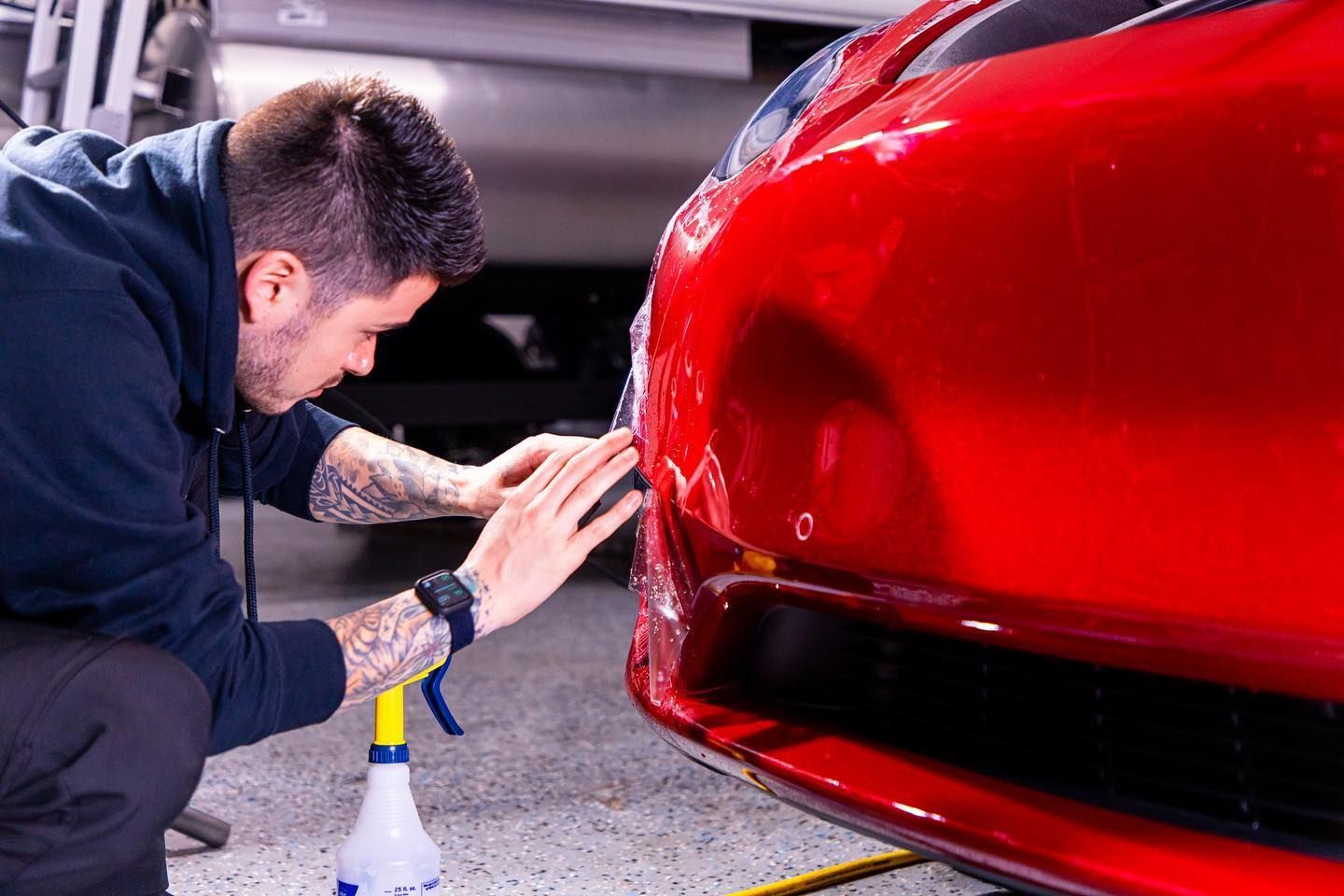Man applying clear film to a red car bumper in a garage.