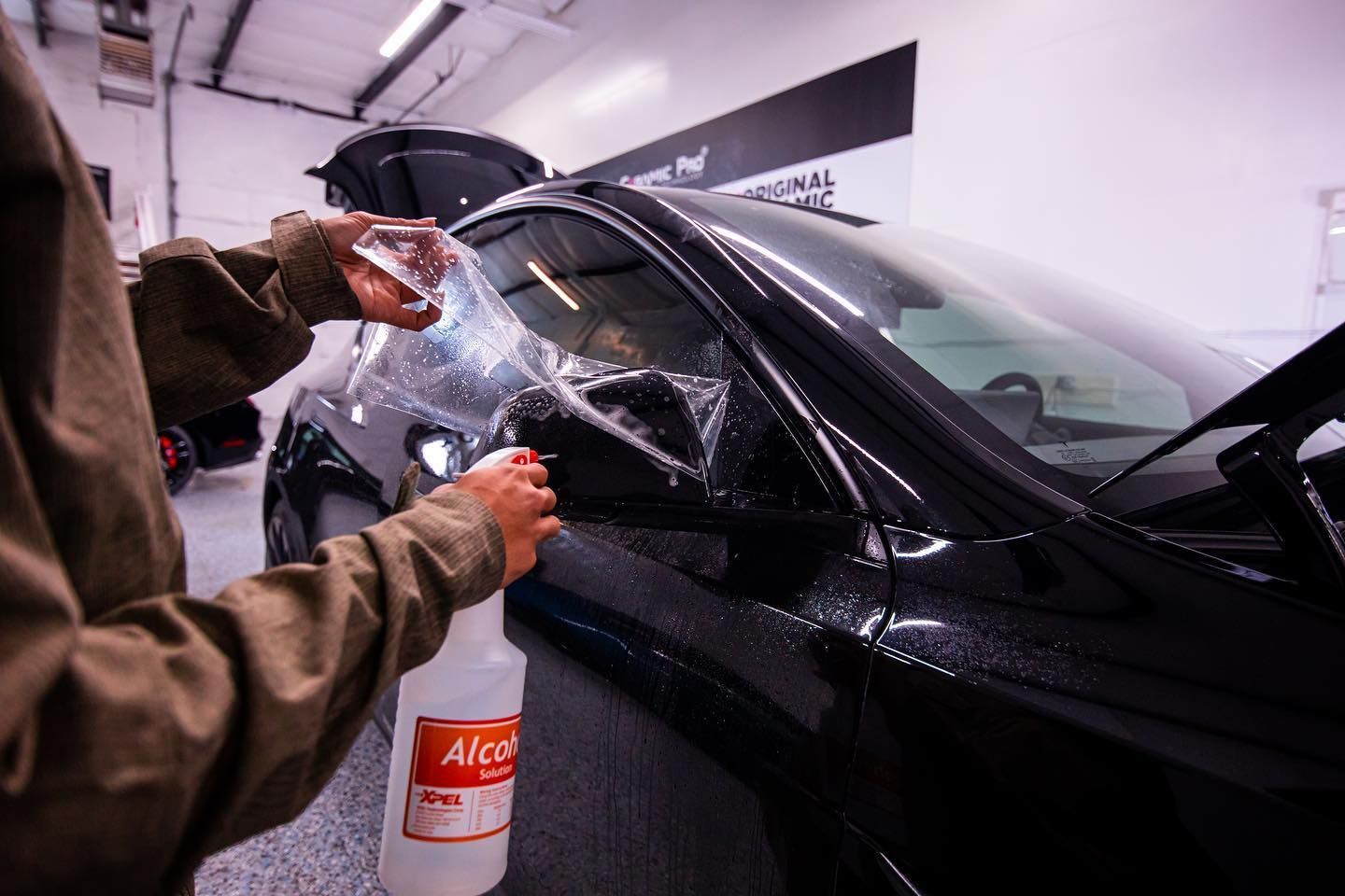 Person sprays a clear liquid on a car's side mirror, preparing to apply a protective film.
