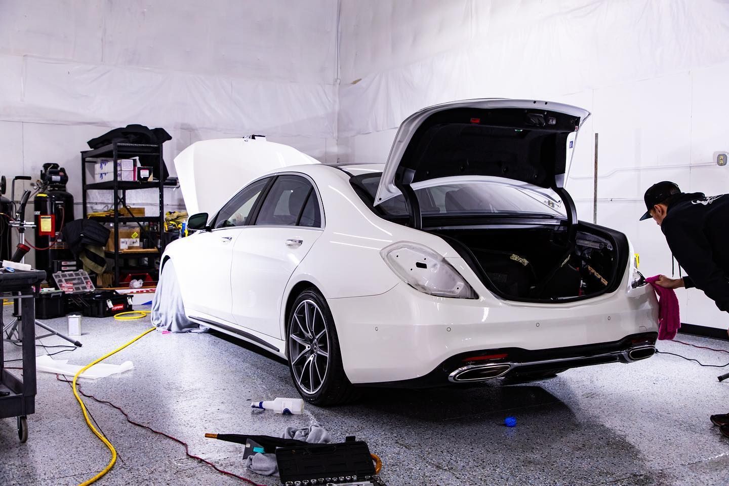 White Mercedes sedan being worked on in a garage; trunk and hood open, worker in frame.