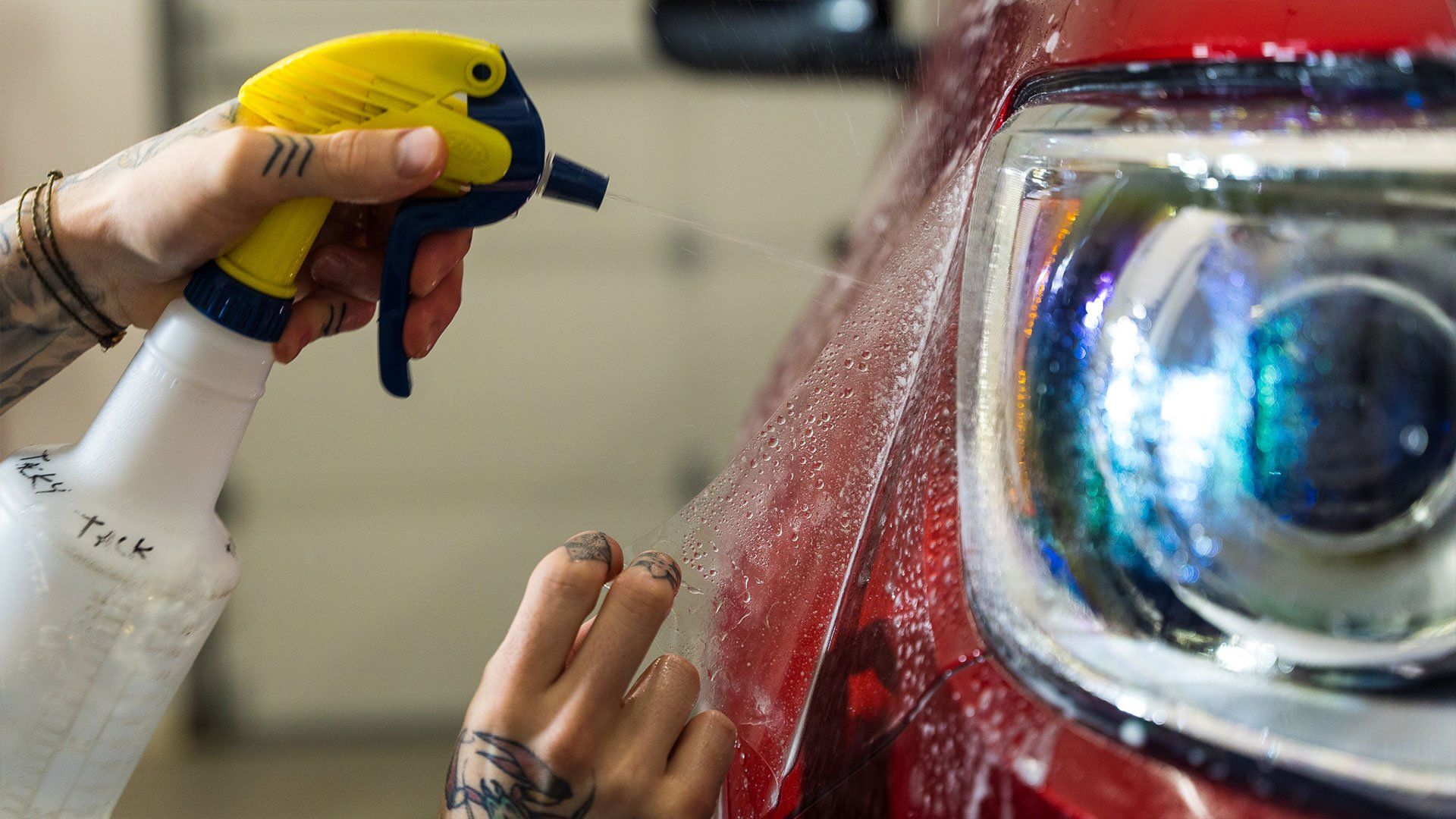 Person applying protective film to a red car headlight with a spray bottle.