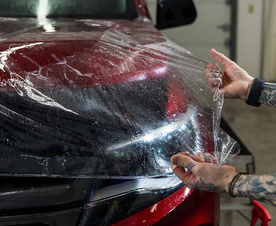 Two hands applying clear film to the red hood of a car.