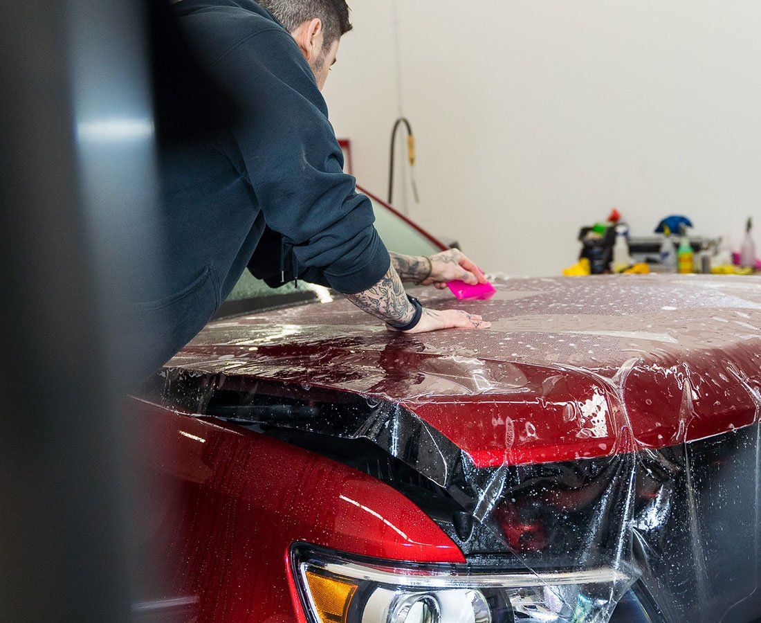 Man applying protective film to a red car hood in a shop.