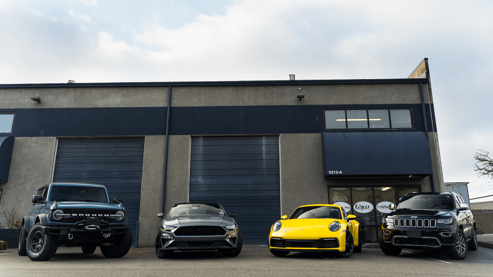 Four cars parked in front of a building: a blue Bronco, gray Mustang, yellow Porsche, and black Jeep.