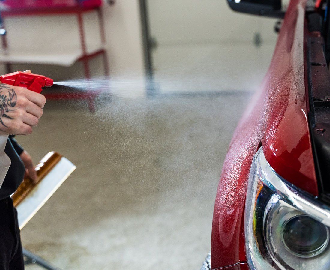 Person spraying water on a red car's fender with a spray bottle, preparing to apply film.