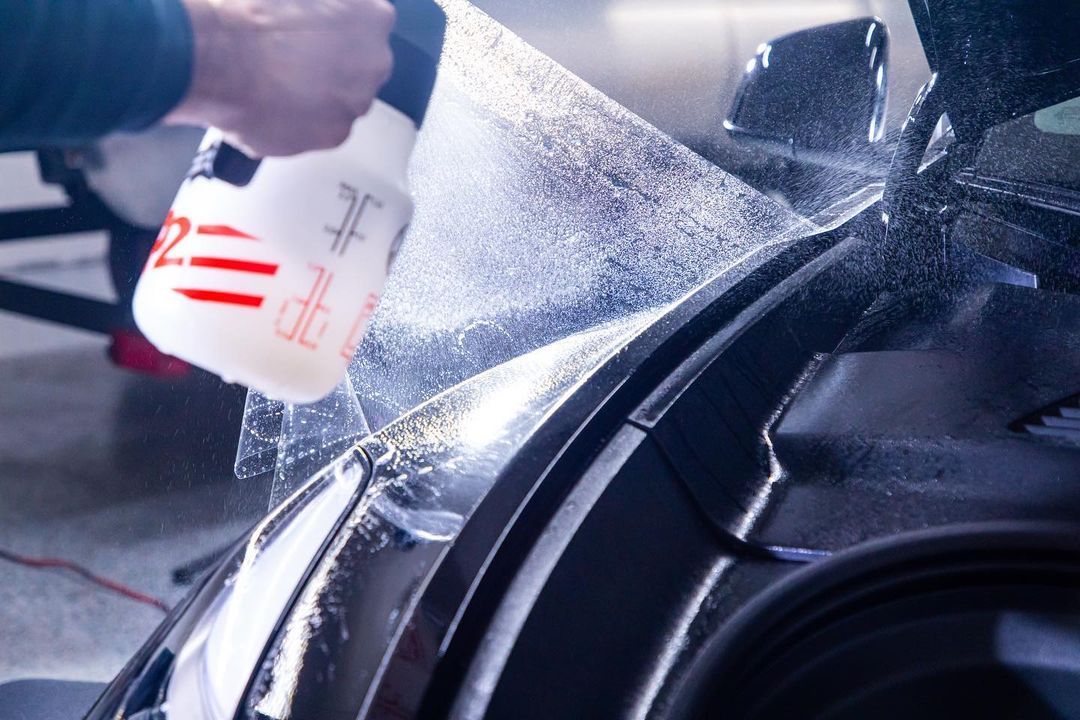 Person spraying a car window with liquid from a white bottle, prepping for window tinting.