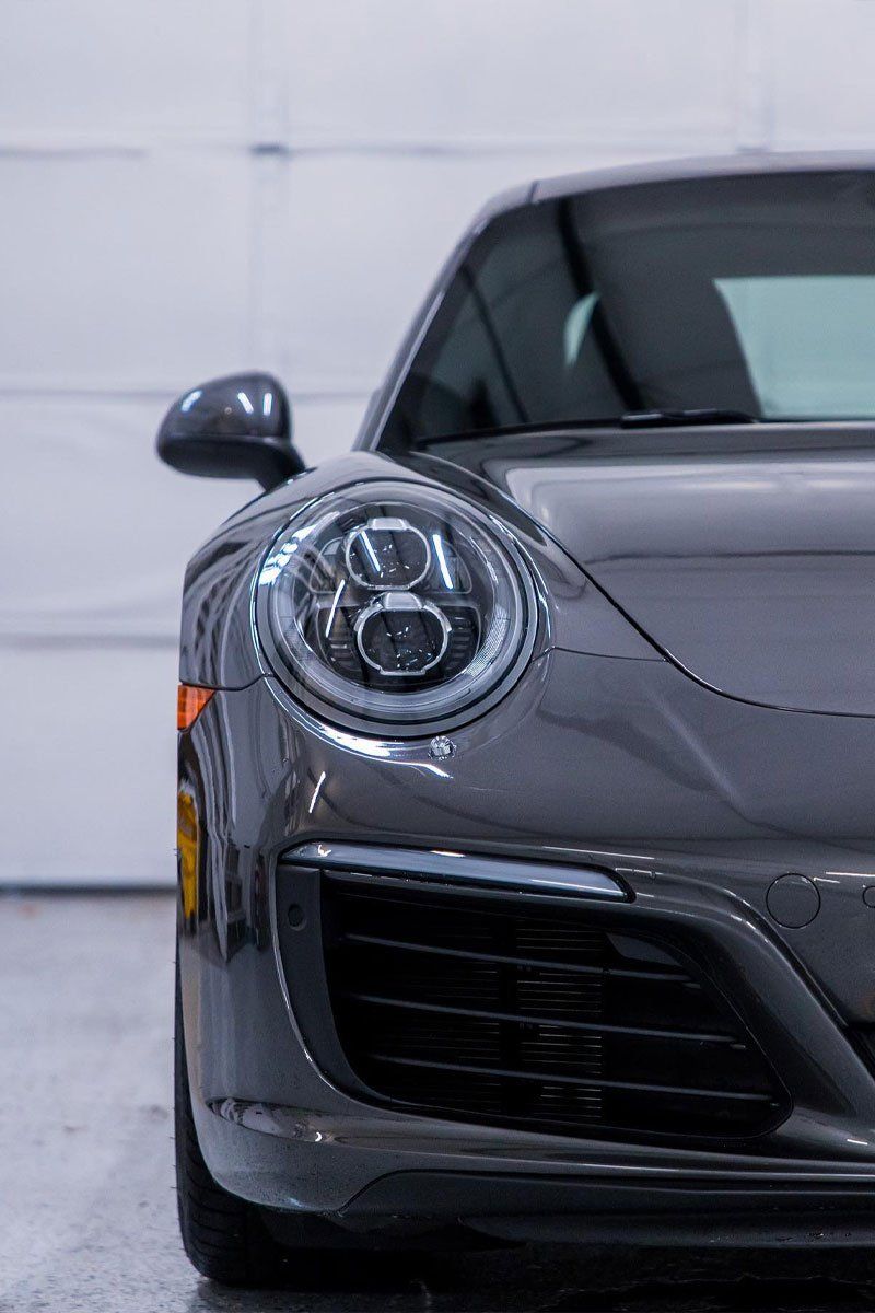 Gray Porsche 911 sports car, front view. In a garage setting, headlights on.