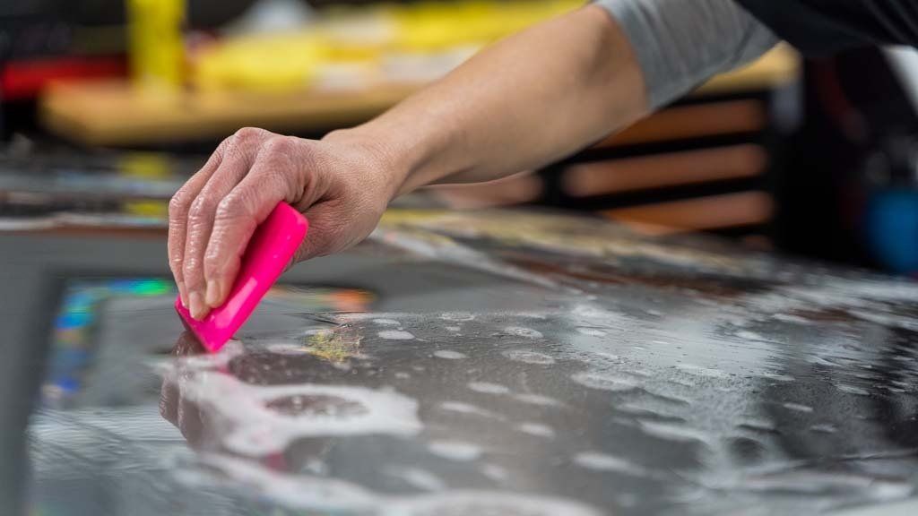Hand using pink squeegee on car's wet surface, possibly applying film in a workshop.