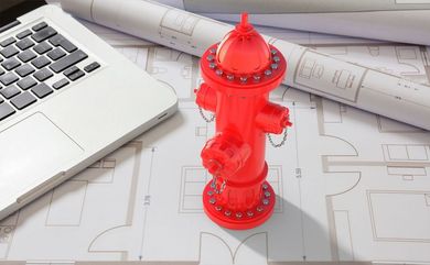Idrante antincendio giocattolo rosso su un progetto architettonico, accanto a un laptop e a dei progetti arrotolati