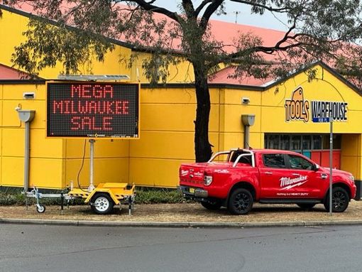A red Milwaukee branded pickup truck parked in front of a yellow Tools Warehouse building with a digital sales sign.